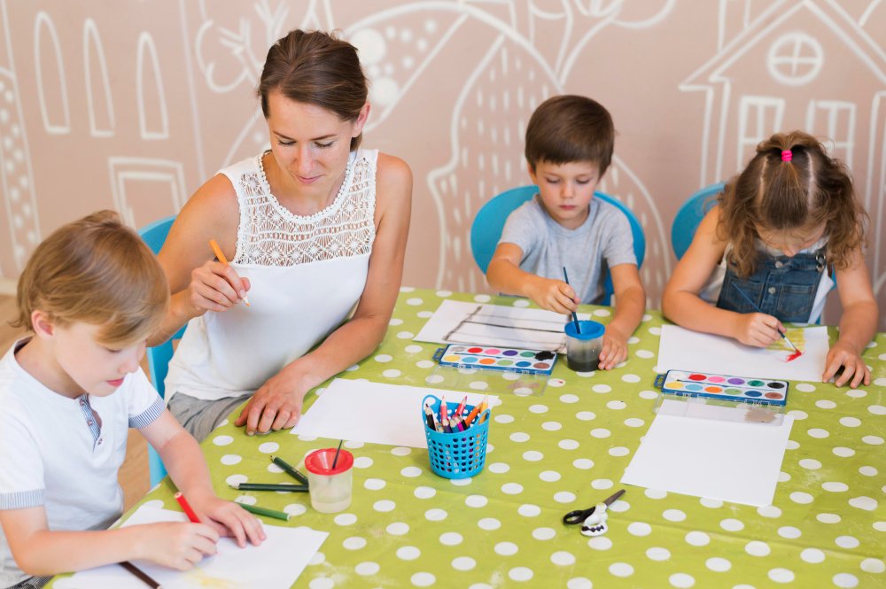 A woman and children sitting together at a table, enjoying a painting session with paper and colorful paints.
