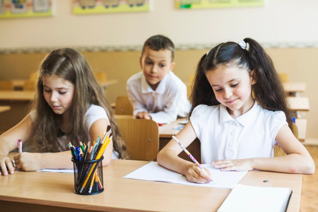 Two girls write in their notebooks at their desks. A boy looks over their shoulders curiously.
