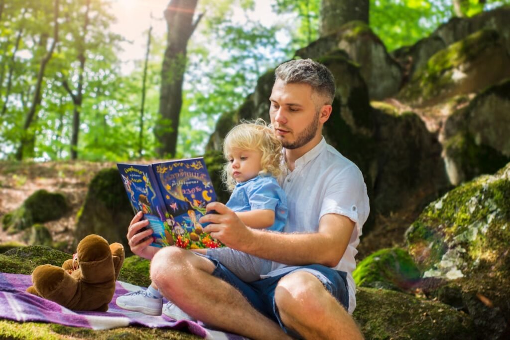 Role of Parents as Partners- a man sitting outdoors on a blanket, reading a storybook to a young child.